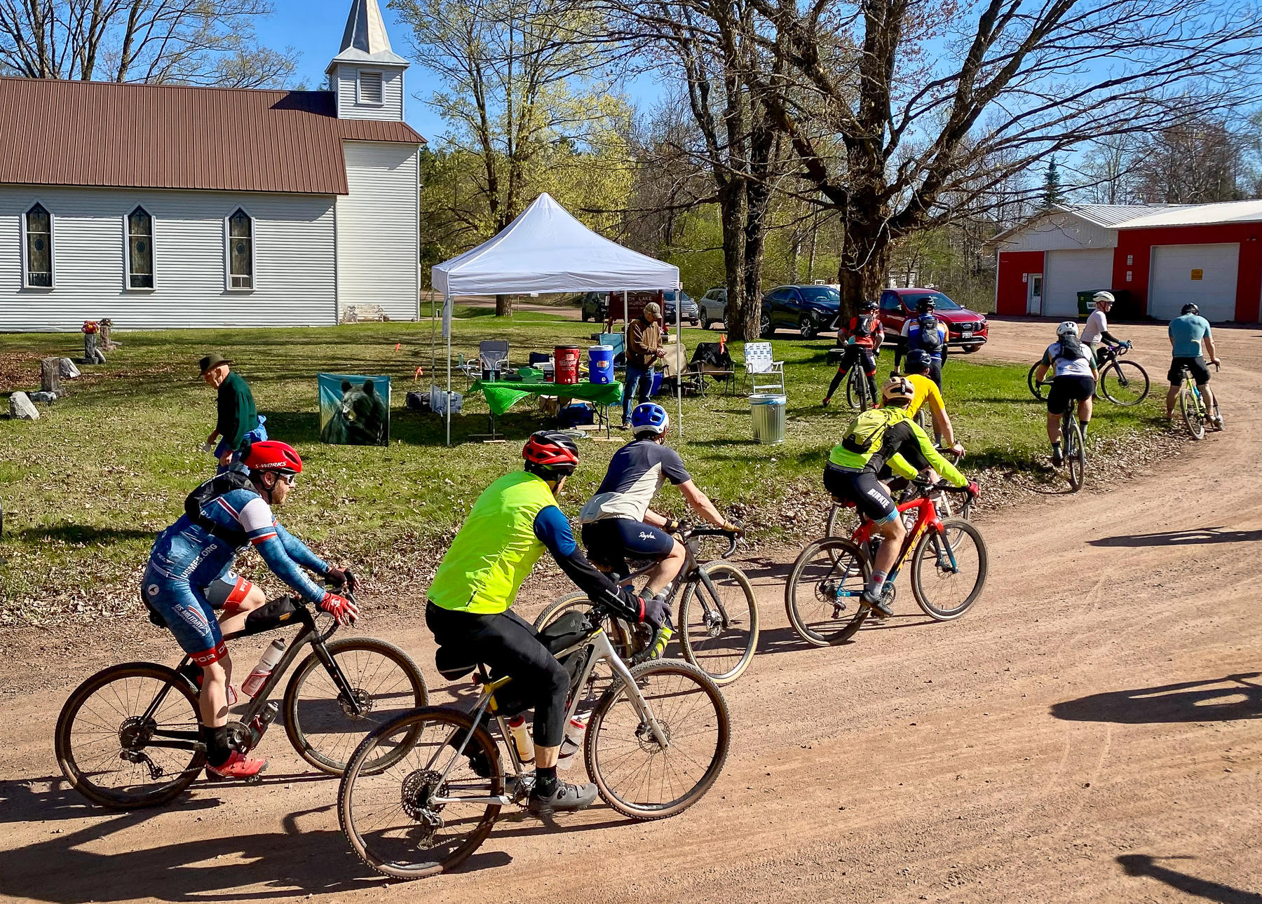 Group of cyclists on a dirt road beside a church, with a white event tent and parked cars in the background.